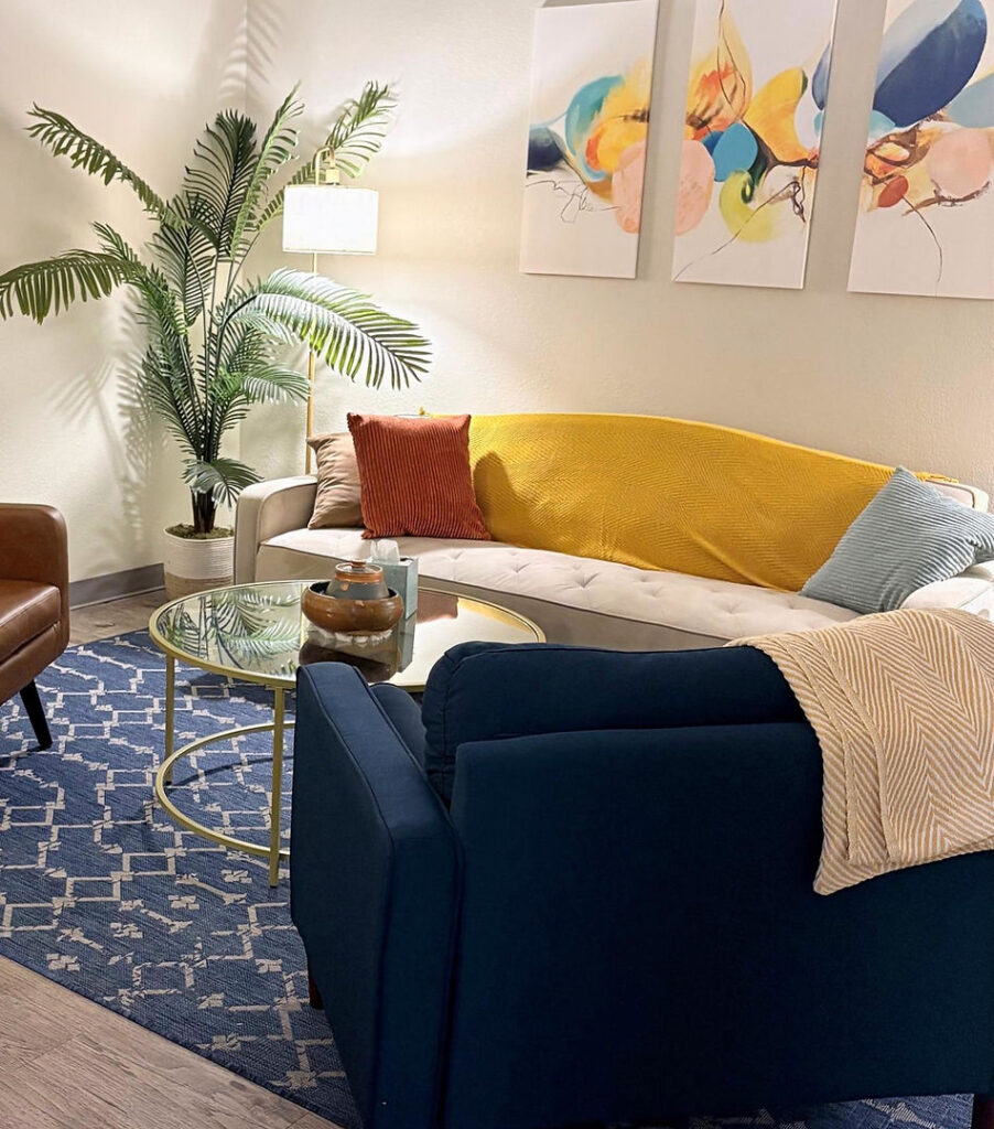 Cozy therapy room with a curved sofa, colorful throw pillows, and a coffee table, featuring a potted plant and abstract wall art, reflecting a warm and inviting atmosphere for mental health support.