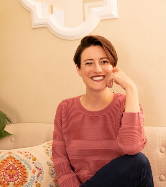 Smiling therapist in a cozy setting, wearing a pink sweater, seated on a couch with a decorative pillow, reflecting a welcoming and personal approach to mental health care.
